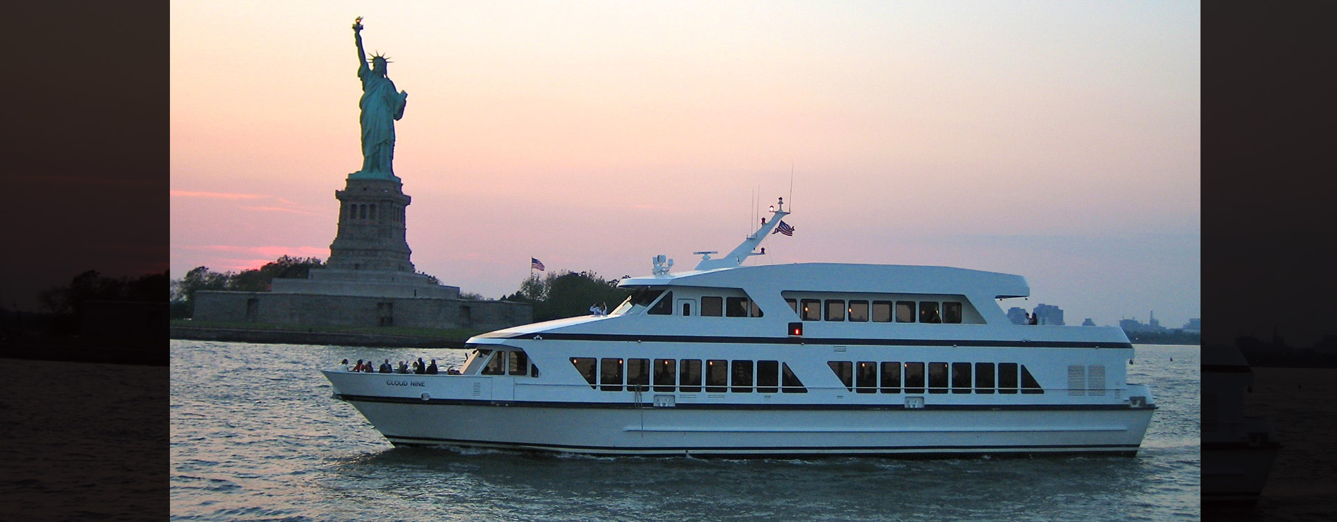 A luxurious yacht sails near the Statue of Liberty at sunset